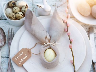  Auf einem Teller liegt ein gekochtes Ei, um das eine Serviette so gewickelt ist, das sie wie Hasenohren aussieht. Daran ist ein Schild befestigt, auf dem "Frohe Ostern" steht.