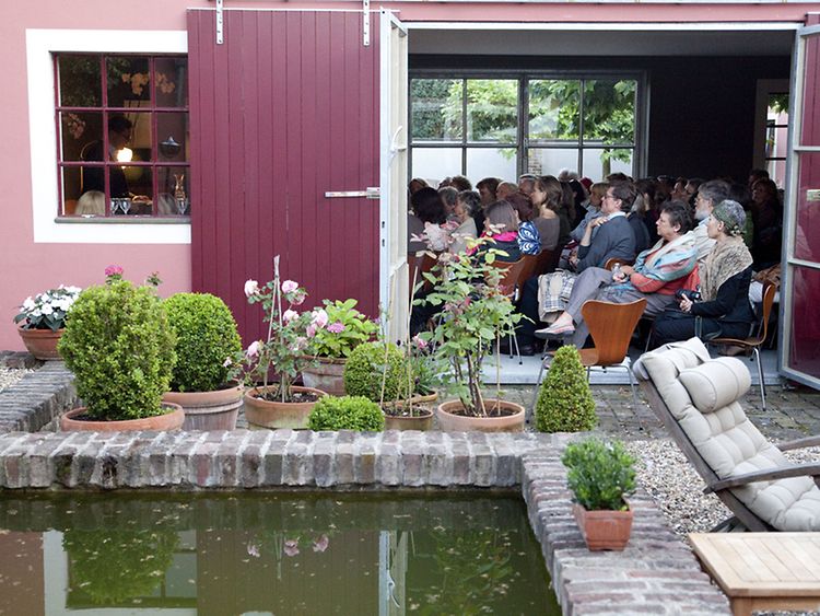  Publikum in einer Scheune lauscht einem Vortrag, im Vordergrund ist ein Garten mit gemauertem Teich zu sehen.