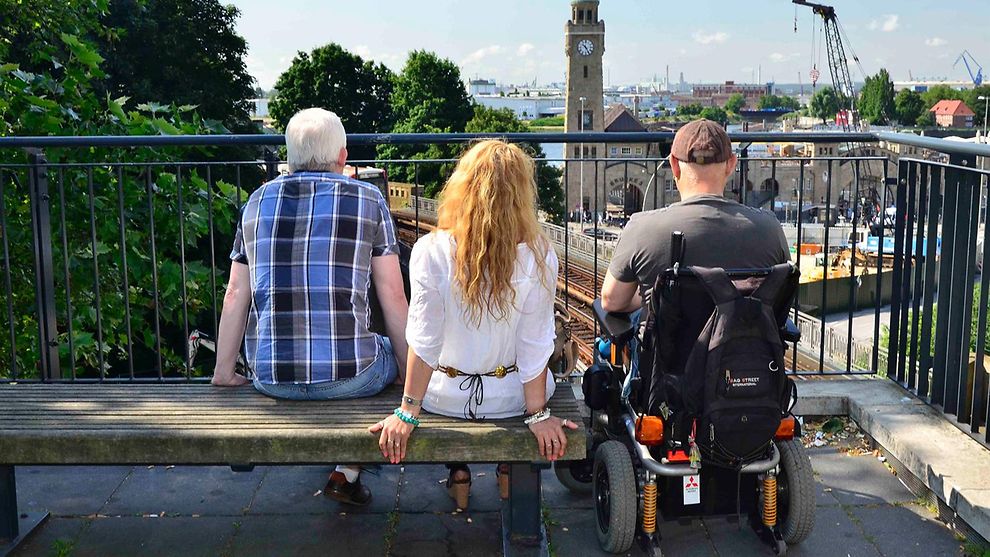 Ein Rollstuhlfahrer und zwei weitere Personen genießen den Blick auf die Hamburger Landungsbrücken