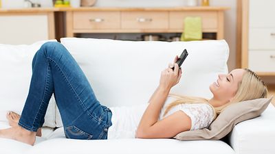  Bild einer jungen Frau, die mit Handy auf einem hellen Sofa liegt
