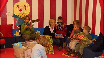  Kleinere Kinder sitzen in einer gemütlichen, bunten Ecke der Bücherhalle und schauen sich Bücher an.