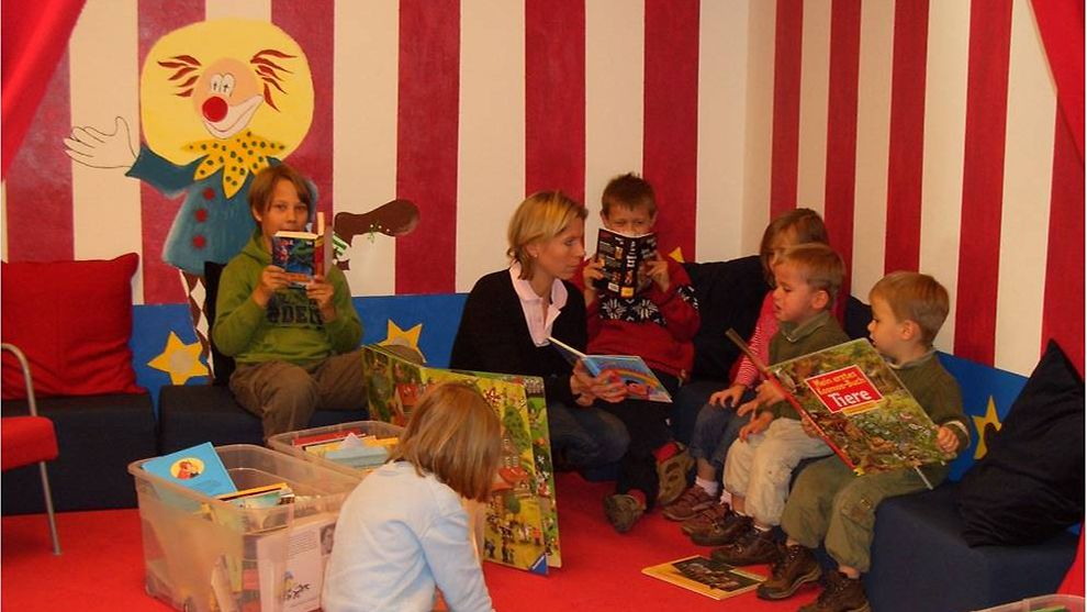 Kleinere Kinder sitzen in einer gemütlichen, bunten Ecke der Bücherhalle und schauen sich Bücher an.