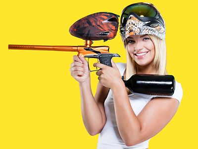  Eine Frau steht vor einem gelben Hintergrund. Sie trägt auf dem Kopf einen Helm mit Brille und in den Hängen hält sie eine Paintball-Maschine.
