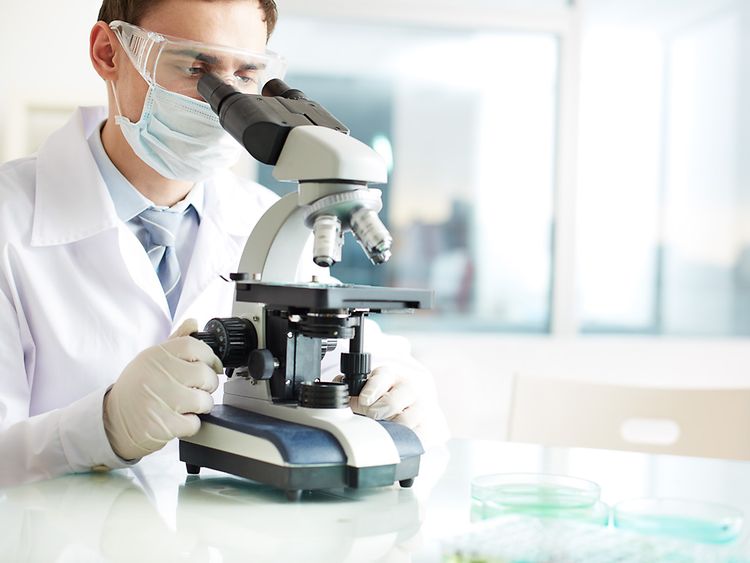  Wissenschaftler mit Schutzbrille und Mundschutz schaut durch ein Mikroskop (Symbolbild)