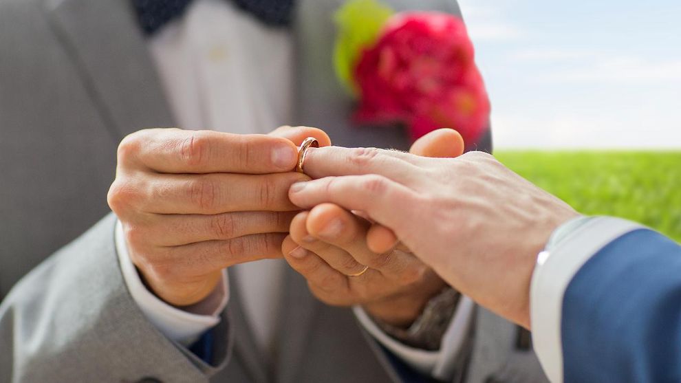 Ein Mann steckt einem anderen Mann einen Ehering auf den Ringfinger der linken Hand. 