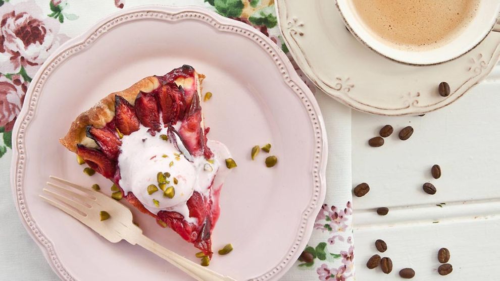  Ein Stück Zwetschgenkuchen auf einem rosa Teller und eine Tasse Kaffee daneben. 