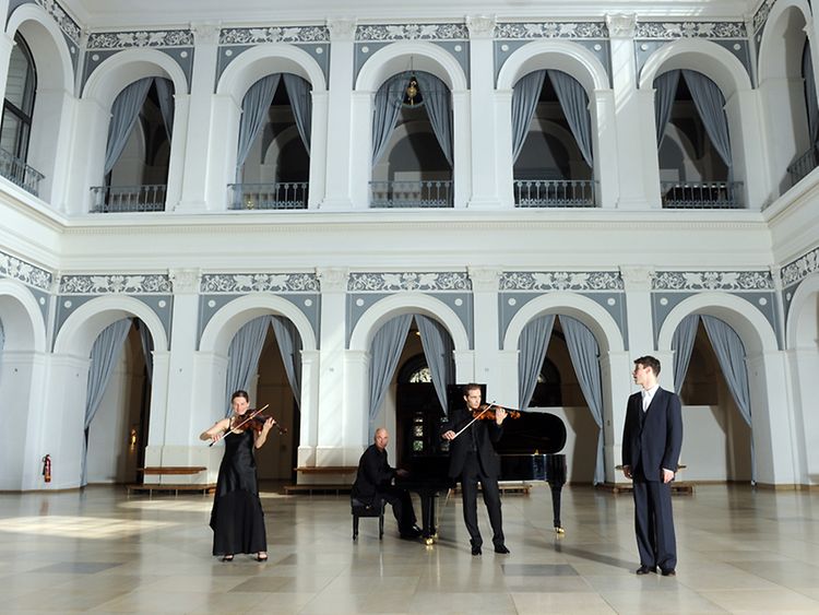  Violinistin, Violinist, Klavierspieler und Sänger musizieren vor zehn verzierten Rundbögen im Börsensaal der Hamburger Handelskammer