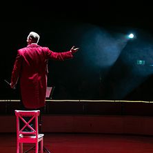  Ein Mann mit einem roten Mantel, von hinten aufgenommen, steht auf einem Stuhl, der auf einer Bühne steht. 