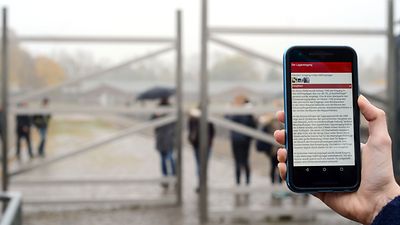  Hand mit Smartphone vor dem Außengelände der KZ-Gedenkstätte Neuengamme