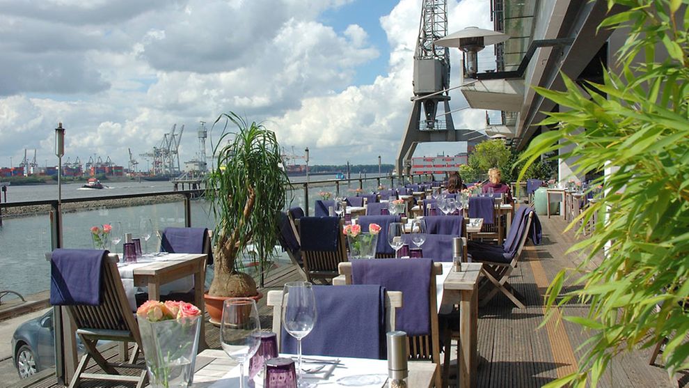  Holzstühle und eingedeckte Tische auf der Außenterrasse des Au Quai; im Hintergrund ist die Elbe zu sehen