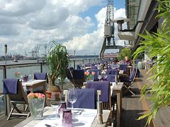  Holzstühle und eingedeckte Tische auf der Außenterrasse des Au Quai; im Hintergrund ist die Elbe zu sehen